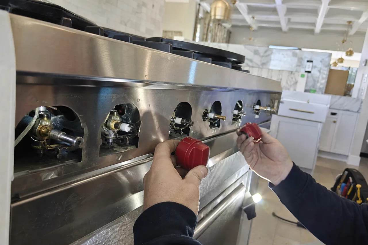 Two hands easing red knobs off a stainless range, the panel open to show brass gas valve stems and white spark switches, white cabinets and marble behind